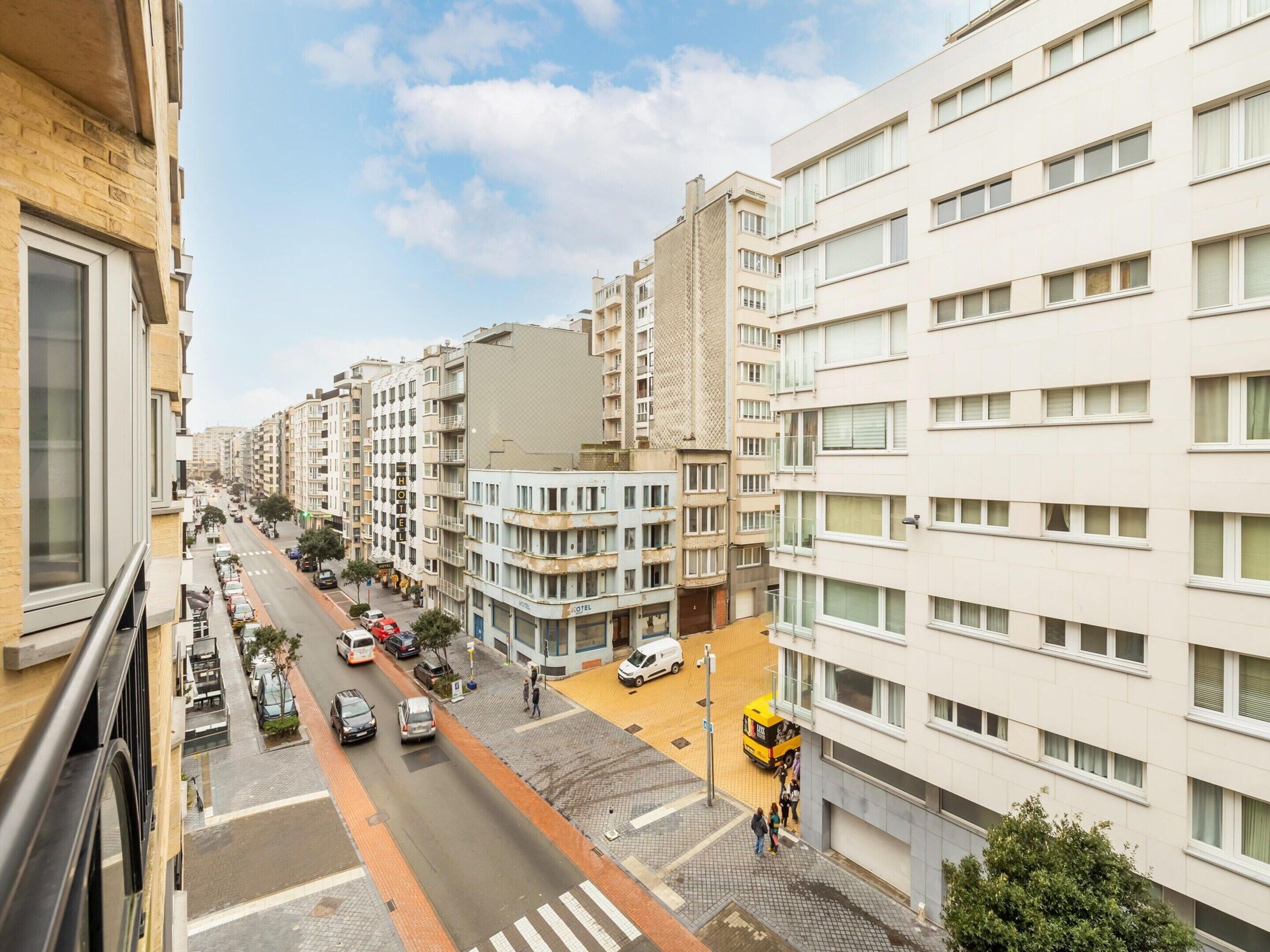 Un hotel de varios pisos con diseño moderno, que cuenta con balcones, un patio central y vistas a la calle con coches estacionados y peatones.