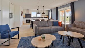 Un salon moderne avec un canapé gris, des fauteuils bleus et des tables rondes en bois. On aperçoit une cuisine à l’arrière-plan, avec un coin repas.