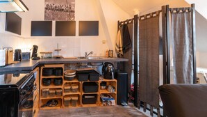 A compact kitchen with a built-in stove, a sink, and a coffee machine. There are wooden shelves with storage containers and a black chair in the corner. - Nice Apartment in Liege With Parking (Liege)