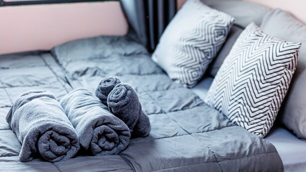 A neatly made bed with grey bedding and rolled towels, a patterned pillow, and a window with curtains. - Baan Mee Suk Ayutthaya (Ayutthaya)