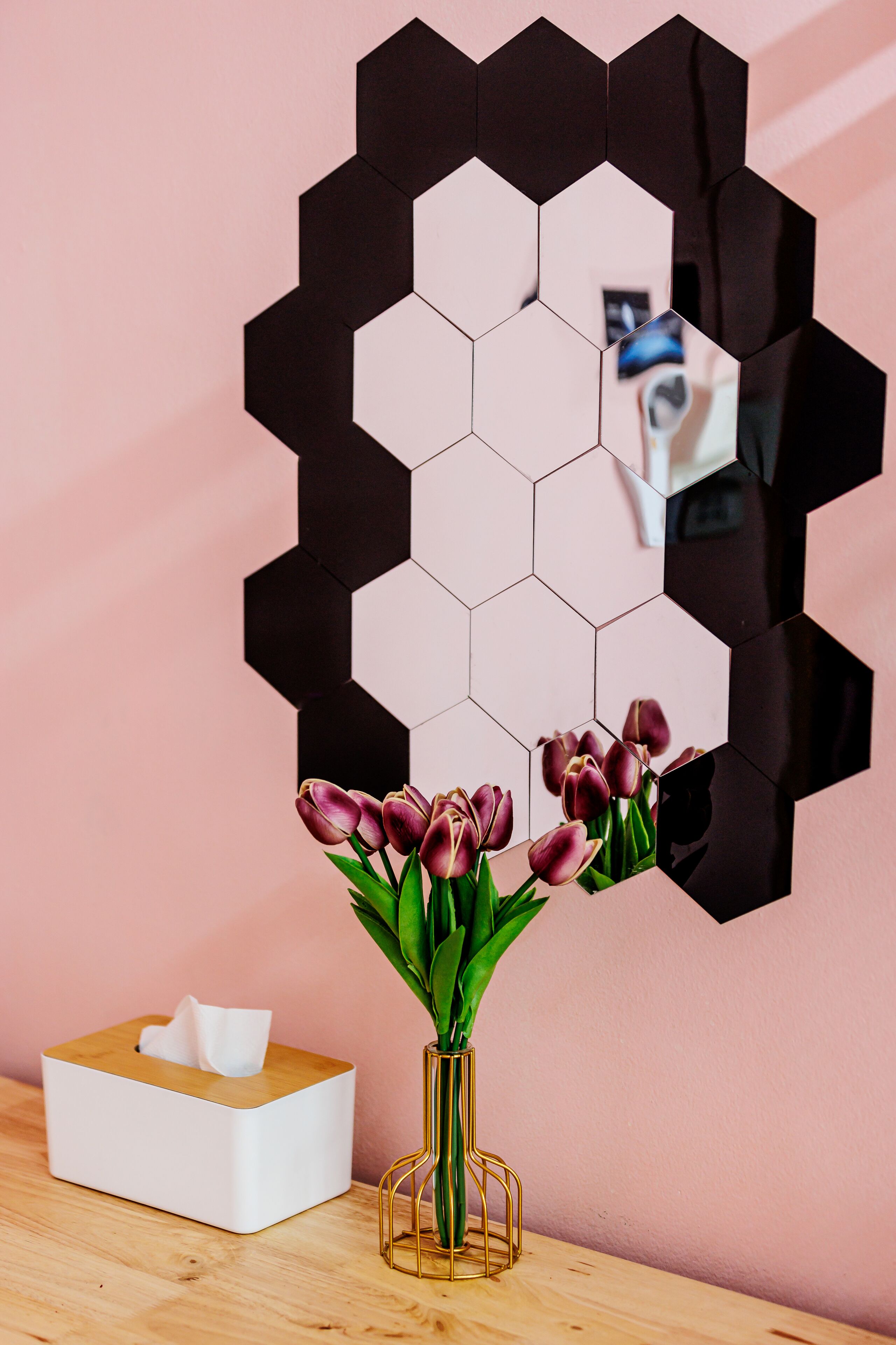 Una habitación con un espejo hexagonal en la pared, una caja de pañuelos y un florero con flores sobre una superficie de madera.