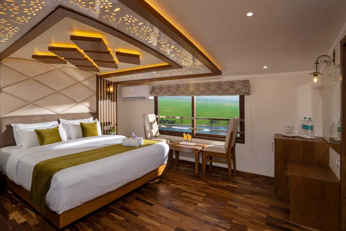 Quarto de hotel com uma cama grande, uma mesinha com dois copos de sumo de laranja, uma cadeira e vista para um campo verde pela janela.