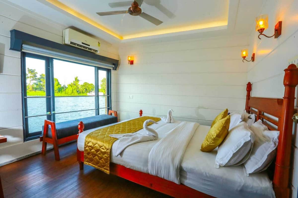 Quarto de hotel com uma cama grande, janela vista para a água, ventilador de teto e luminárias na parede.