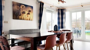A dining area with a wooden table and chairs, a painting on the wall, and large windows with curtains. - Bungalow With Sauna & Nature (Earnewald)