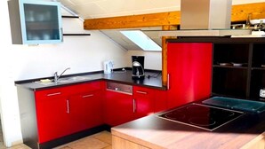 A compact kitchen with red cabinets, a built-in dishwasher, a coffee maker, and a black induction cooktop. - Junkerath Romantic Garden Stay (Juenkerath)