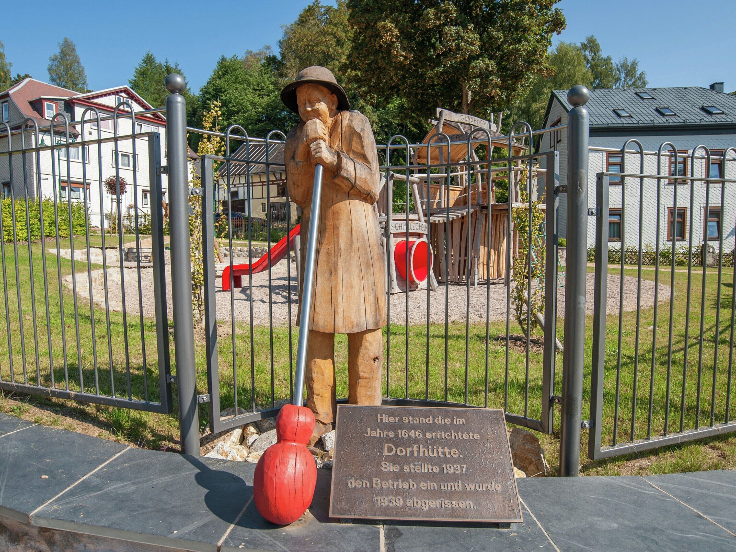 A wooden statue of a man with a shovel and a red ball, standing in front of a playground with a slide and a swing set.