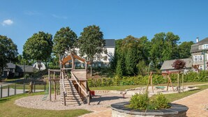 Um parquinho com uma estrutura de escalada de madeira e balanços, rodeado por um jardim paisagístico e edifícios residenciais ao fundo.