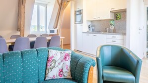 Ein gemütliches Wohnzimmer mit einem türkisfarbenen Sofa, Holzboden und Blick auf eine Küche mit weißen Schränken und einer Mikrowelle.