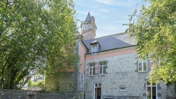 Ein Steinhaus mit einem Turm, einem Rasenstück und einem Metall-Stuhltisch.