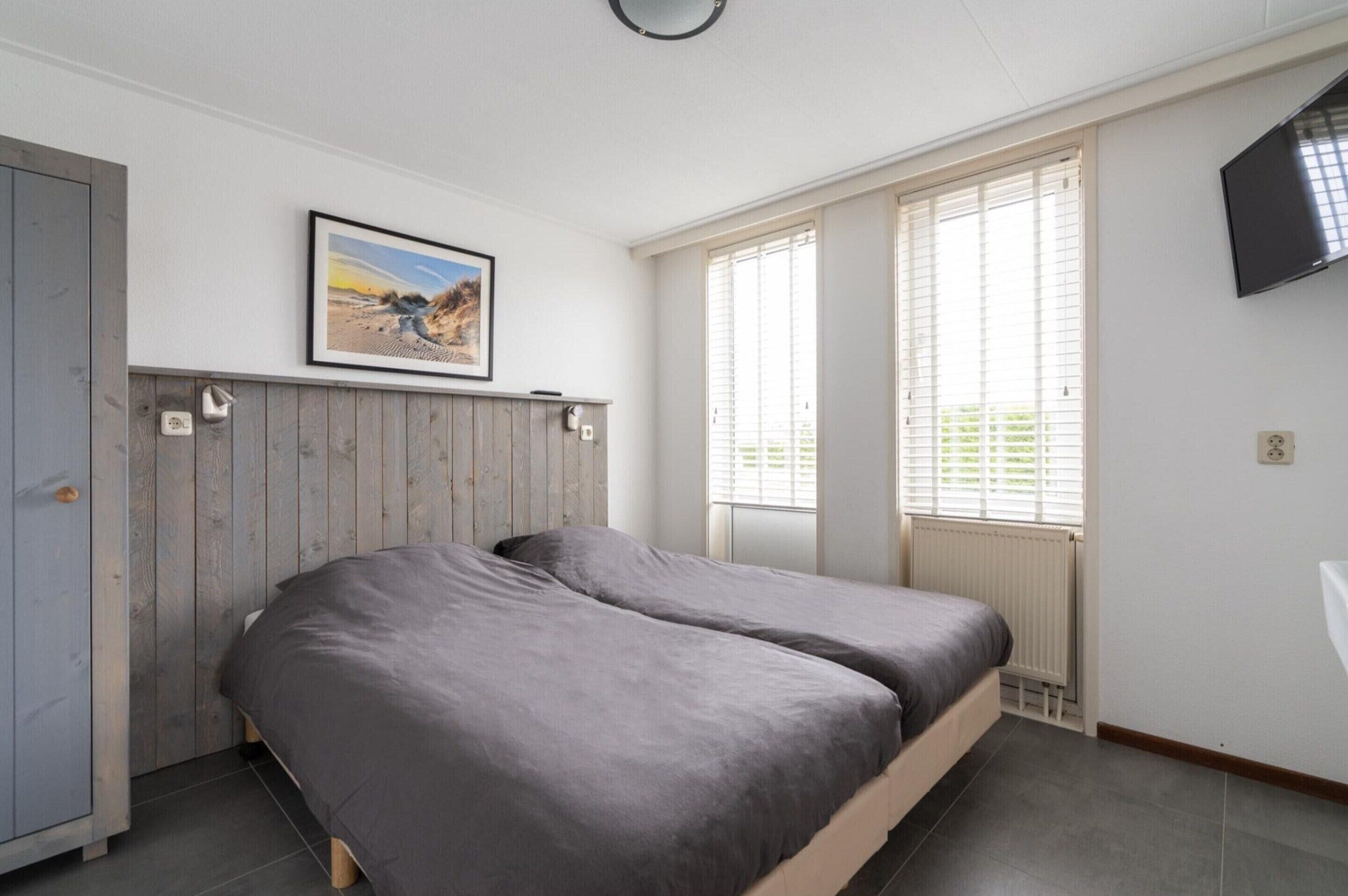 A bedroom with a wooden headboard, a bed with grey bedding, a mounted TV, and a window with blinds.