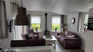 A modern living room with a sofa, chairs, a coffee table, and a dining table. There are windows with curtains, a wall-mounted TV, and a clock. - Cosy Holiday Home in Appelscha With Garden (Appelscha)