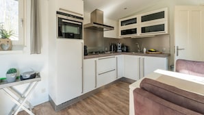 A modern kitchen with white cabinets, a built-in oven, and a stove. There is a window with curtains, a small table with a plant, and a pink chair. - Cosy Holiday Home in Appelscha With Garden (Appelscha)
