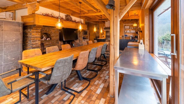 A long wooden dining table with cushioned chairs, a stone fireplace, and a large TV screen. - Home in Heart of Belgian Nature (Vresse-sur-Semois)