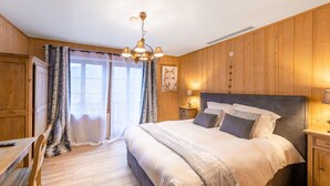 A bedroom with a wooden floor, a bed with white and gray bedding, a nightstand with a lamp, and a window with curtains. - Home in Heart of Belgian Nature (Vresse-sur-Semois)