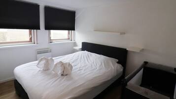 A bedroom with a bed, two windows with blinds, a radiator, and a TV stand.