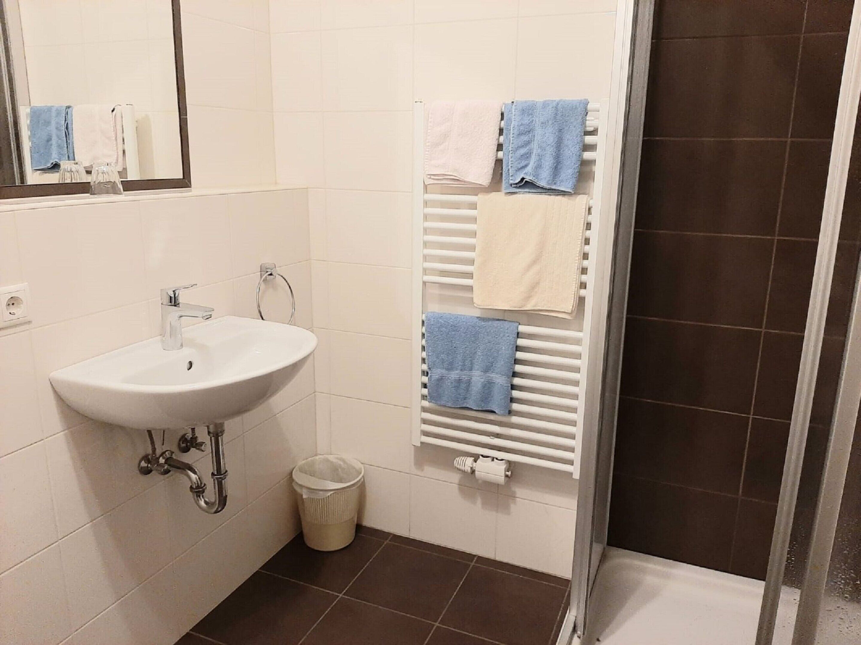 A bathroom with a white sink, a mirror, a towel rack with multiple towels, a waste bin, and a shower area.