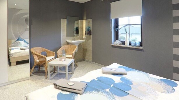 A modern bedroom with a large bed, a small table, two wicker chairs, a bathroom with a sink and mirror, and a window with a view. - Holiday Home in Gonnersdorf With Sauna (Gonnersdorf)
