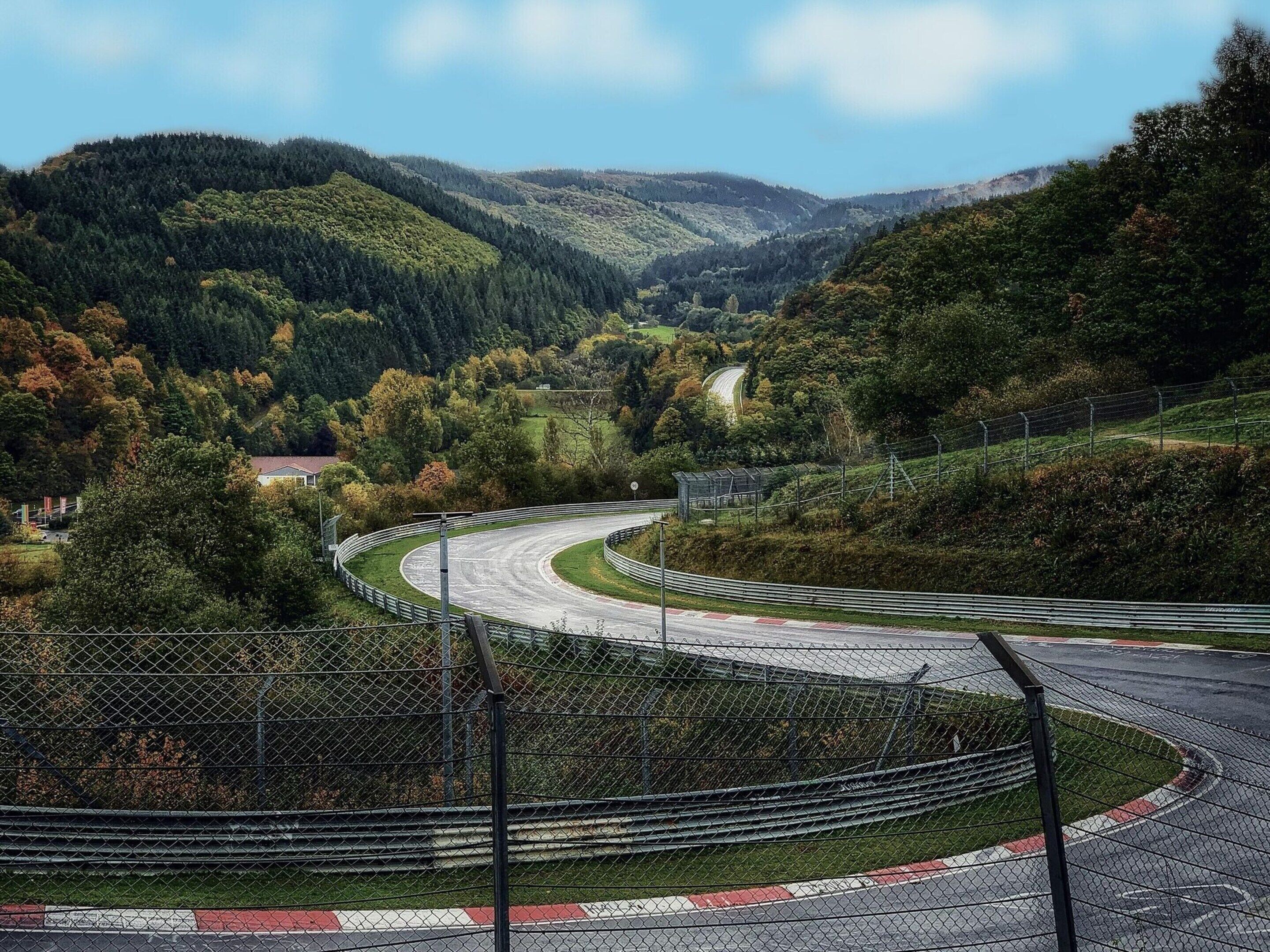 A winding road with a fence and guardrails, surrounded by lush green hills and trees.