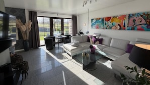 A modern living room with a sofa, a glass coffee table, and a TV. There are large windows and a painting on the wall. - Holiday Home in Gonnersdorf With Sauna (Gonnersdorf)