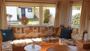 A cozy RV interior with a sectional sofa, a small round table, and large windows offering a view of a house and greenery. - Nice Holiday Home in Appelscha With Garden (Appelscha)