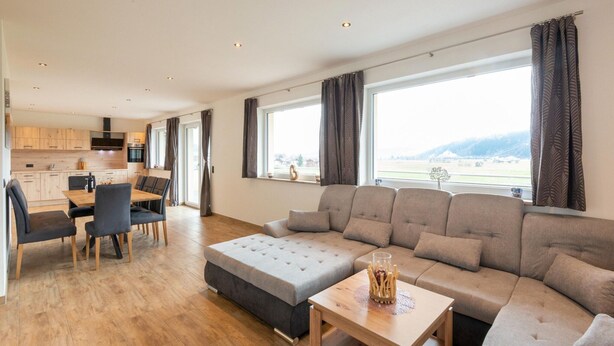 Ein modernes Wohnzimmer mit einer Eckcouch, einem Esstisch mit Stühlen und einer Küche im Hintergrund.