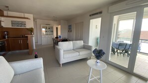 A spacious living area with a white sofa, a small round table with a vase, and a kitchen area with wooden cabinets and a dining table.