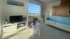 A living room with a TV, a sofa, a coffee table, and a balcony with a view of the sea.