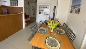 A dining area with a wooden table set for four, surrounded by chairs. There is a kitchen area to the left with a coffee machine and a counter. A dining table with a vase of flowers is in the center.