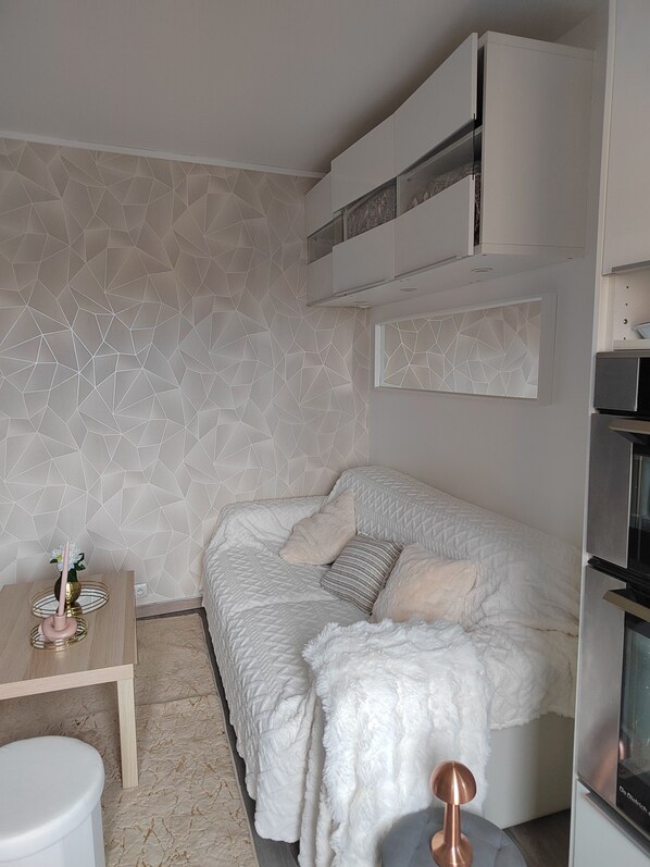 A modern bedroom with a geometric-patterned wall, a bed with white bedding, a small wooden nightstand, and a built-in oven. - Studio near La Défense, close to amenities, close to streetcar and bus transport. (Colombes)
