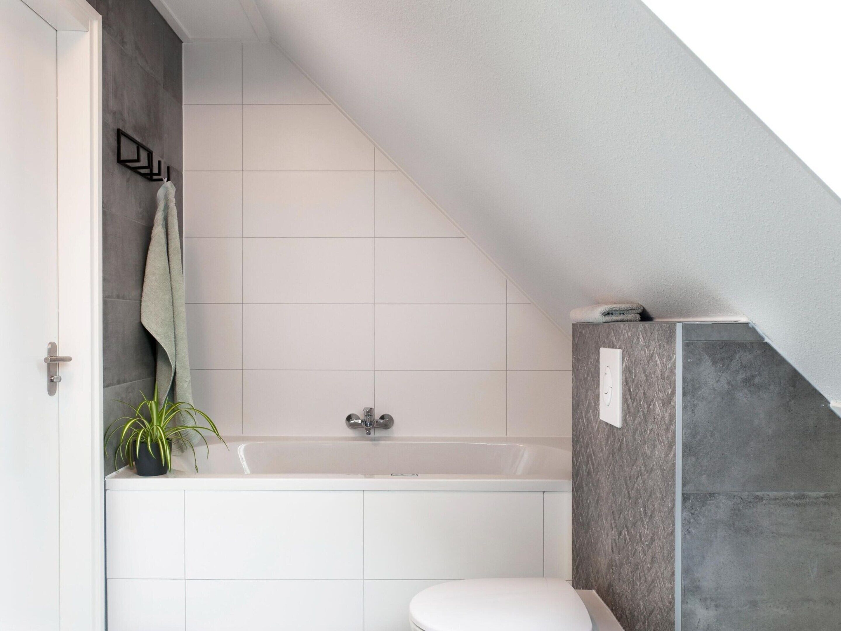 A modern bathroom with a bathtub, white tiles, a towel rack, and a potted plant.