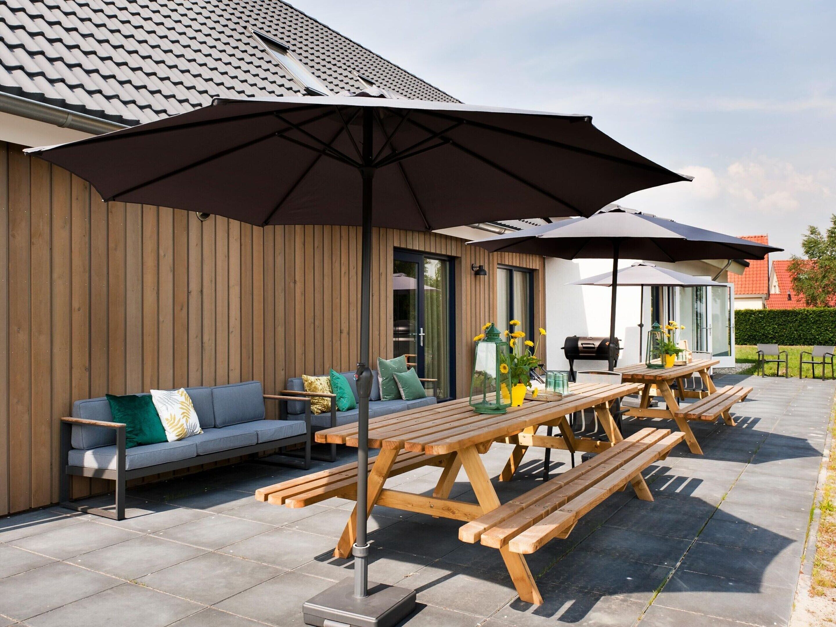 An outdoor patio with wooden benches and a large umbrella, a sofa with cushions, and a grill.