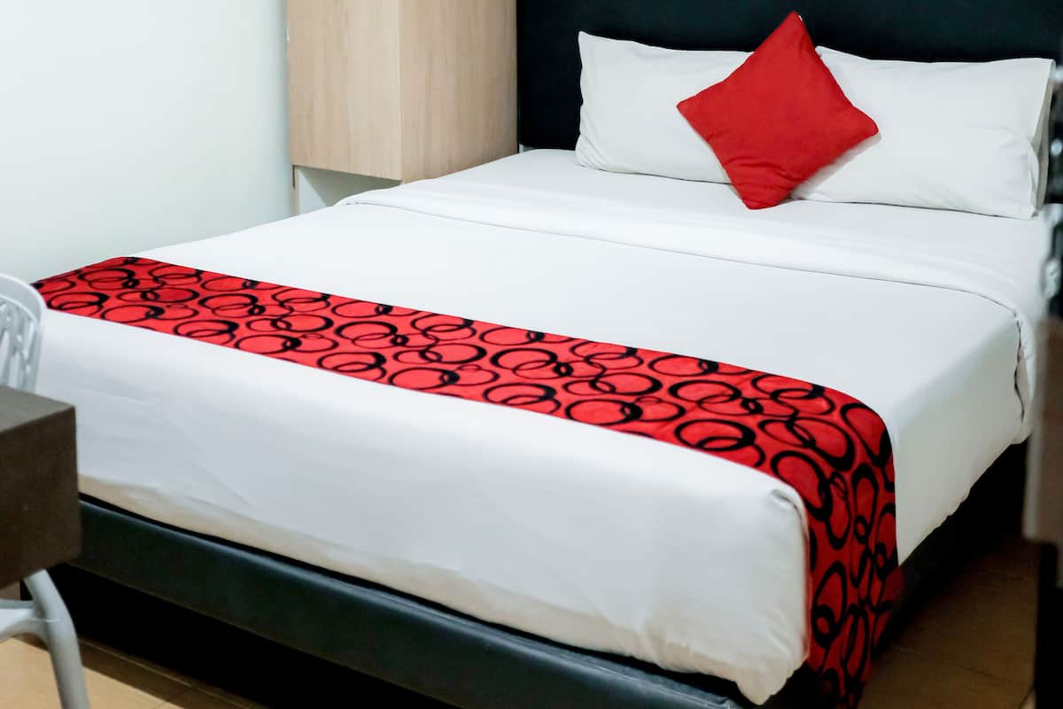 Uma cama de solteiro com colcha branca e vermelha, um guarda-roupa de madeira e uma cabeceira preta.