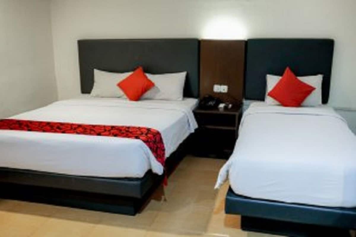 Quarto de hotel com duas camas de solteiro, cada uma com cabeceira escura, roupa de cama branca e um travesseiro vermelho decorativo.