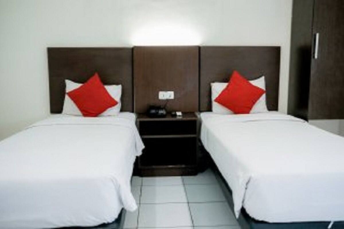 Quarto de hotel com duas camas de solteiro, cada uma com roupa de cama branca e almofadas vermelhas. Há uma mesa de cabeceira entre as camas, com um telefone e uma pequena mesa. O quarto possui uma cabeceira de madeira escura e uma parede branca.