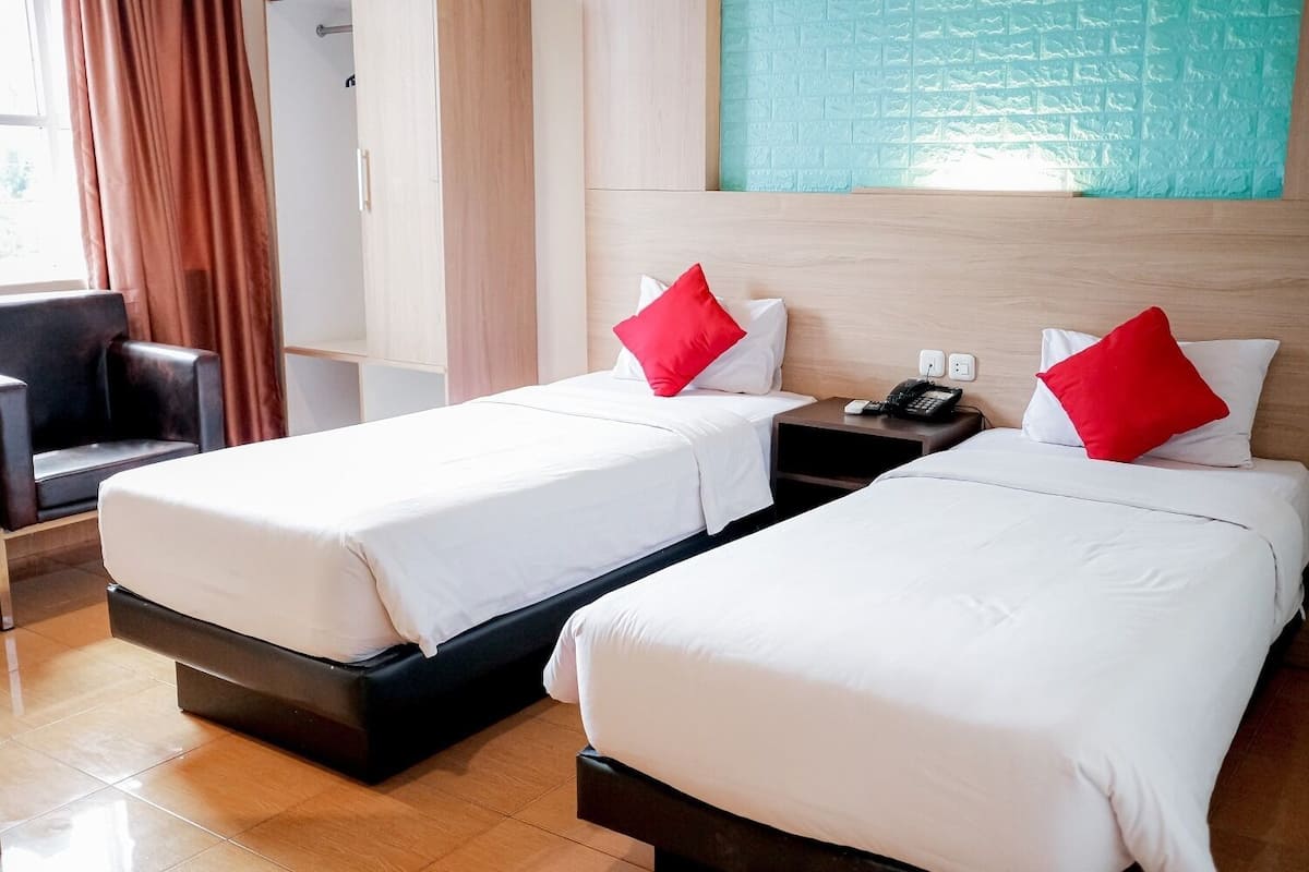 Quarto de hotel com duas camas, cada uma com roupa de cama branca e almofadas vermelhas, uma mesa de cabeceira com telefone, e vista para uma parede azul com textura.