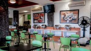 A restaurant with green chairs and tables, a counter with food items, and a fan. - WINER HOTEL PALEMBANG (Palembang)