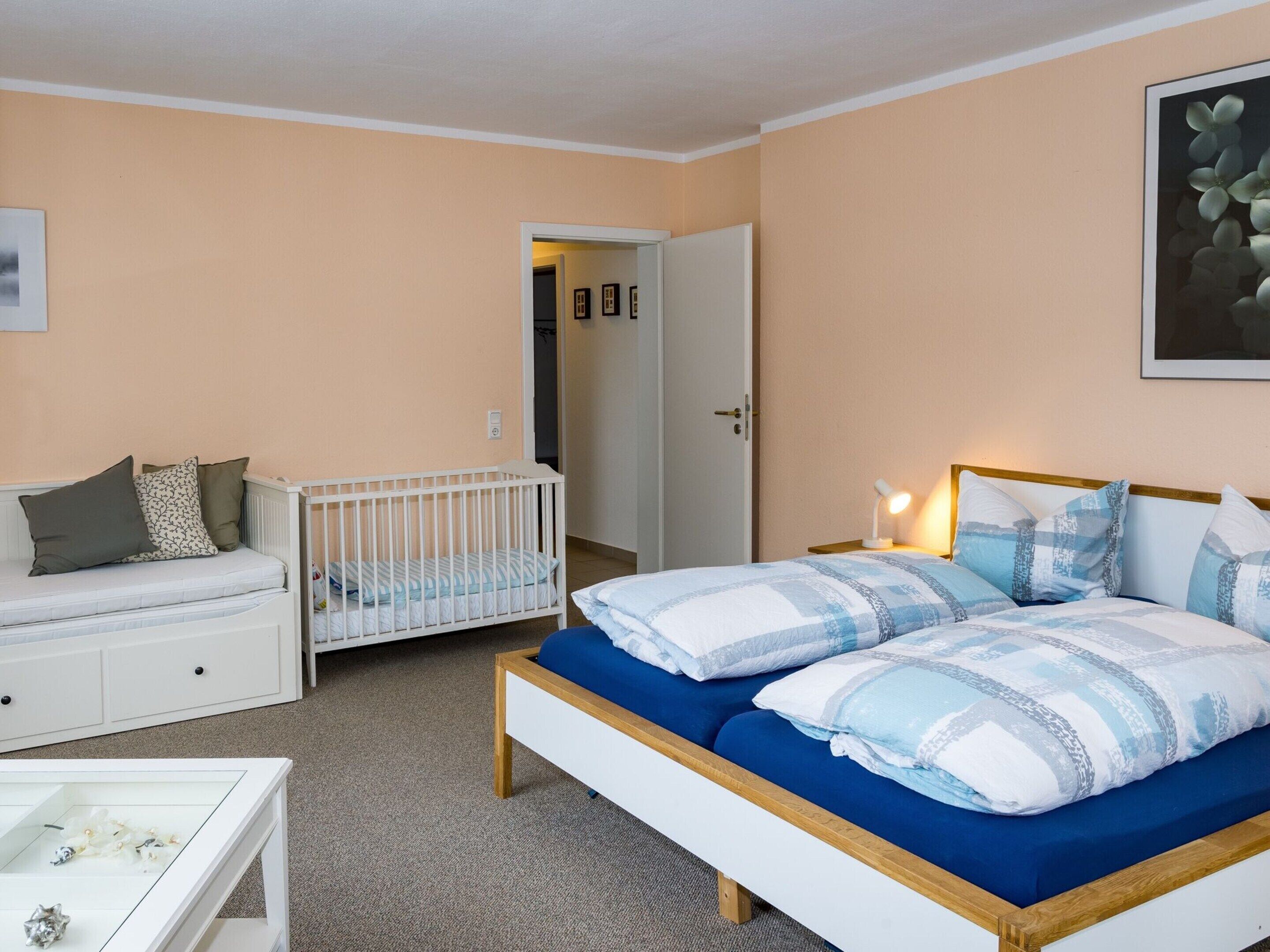 A bedroom with a crib, a bed with blue bedding, a white dresser, and a bedside table with a lamp.