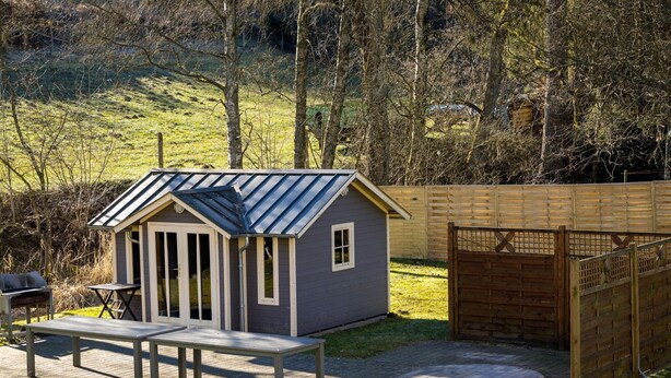 A small, single-story wooden structure with a sloped roof, a patio with a table and benches, and a fenced area. - Ferienwohnung im Sauerland (Erndtebrück)