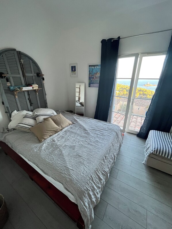 Ein Schlafzimmer mit einem Bett, einem kleinen Nachttisch, einem Spiegel und Meerblick durch ein Fenster.