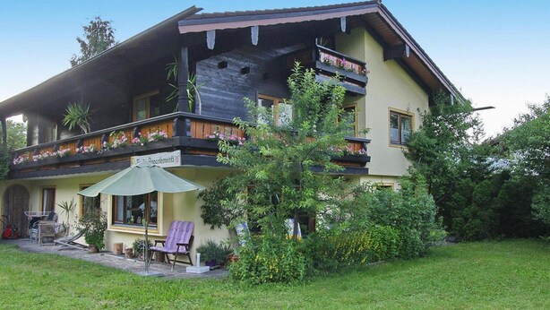 Una casa de dos pisos con un balcón, un patio con muebles de exterior y un jardín.