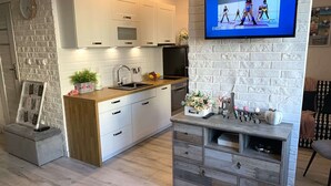 A modern kitchen with white cabinetry, a built-in oven, and a flat-screen TV mounted on a brick wall. - Apartment in Gdynia Near Baltic Beach (Gdańsk)