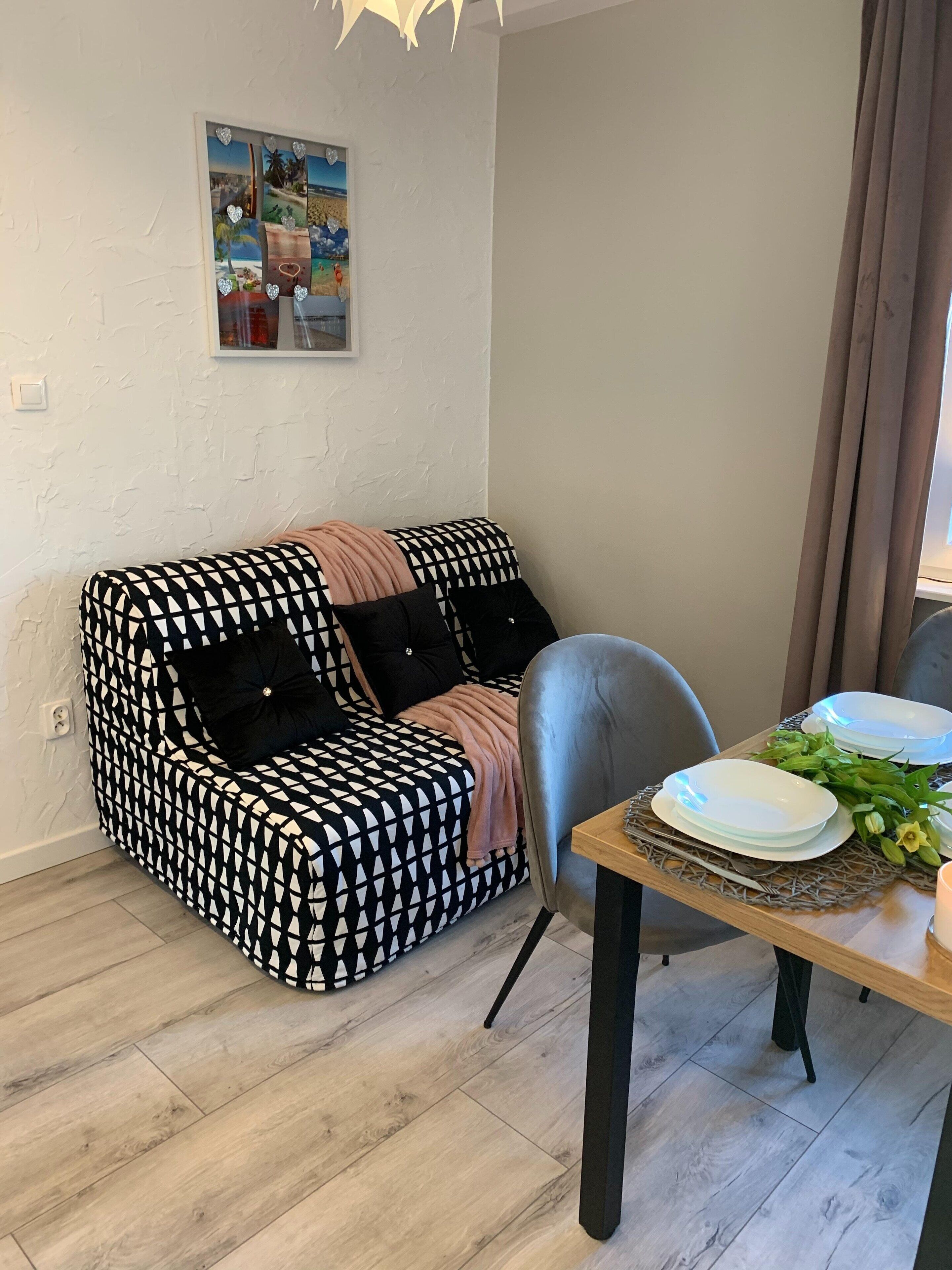 Ett modernt vardagsrum med en mönstrad soffa, ett matbord med tallrikar och bestick samt en dekorativ väggdekoration.