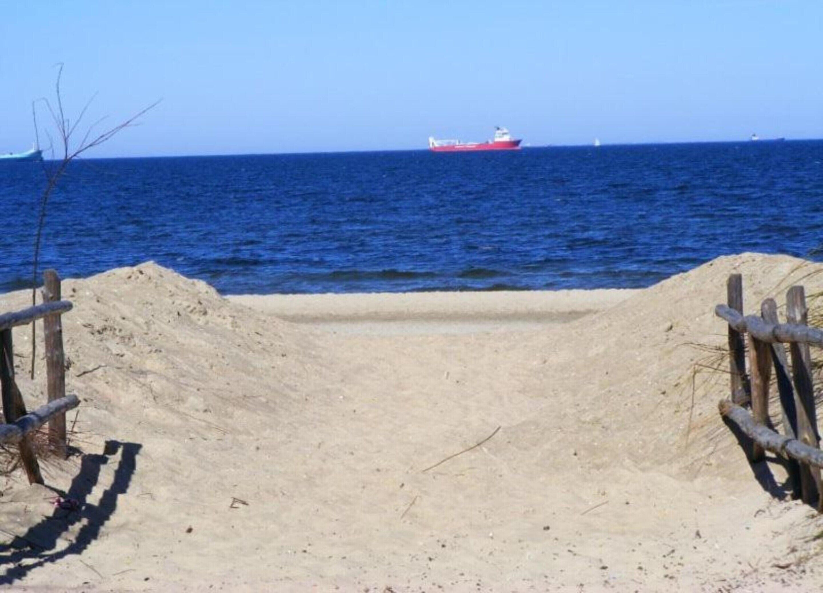 A sandy beach with a wooden fence and a red ship in the distance.