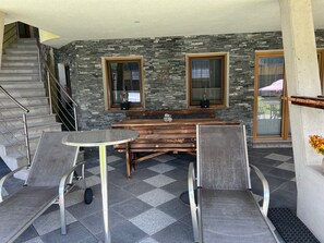 Un área de patio techada con un muro de piedra, un banco de madera, dos sillas de metal y una mesa redonda pequeña.