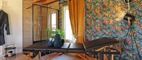 A room with a massage table, a glass shower enclosure, and a tropical wallpaper.