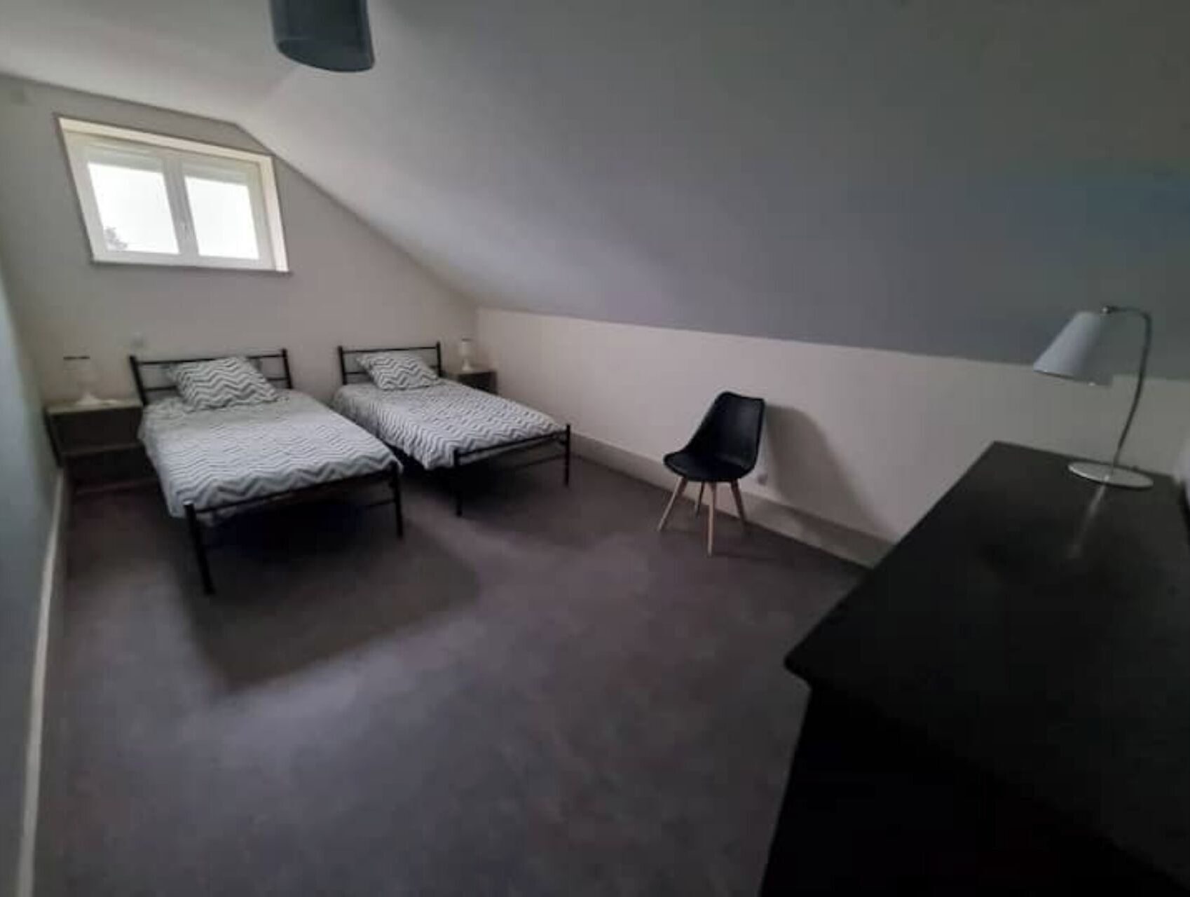 A small, dimly lit room with two single beds, a chair, a desk, and a window.