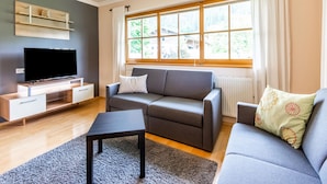 Ein modernes Wohnzimmer mit Flachbildfernseher, grauen Sofas, einem Couchtisch und einem großen Fenster mit Vorhängen.
