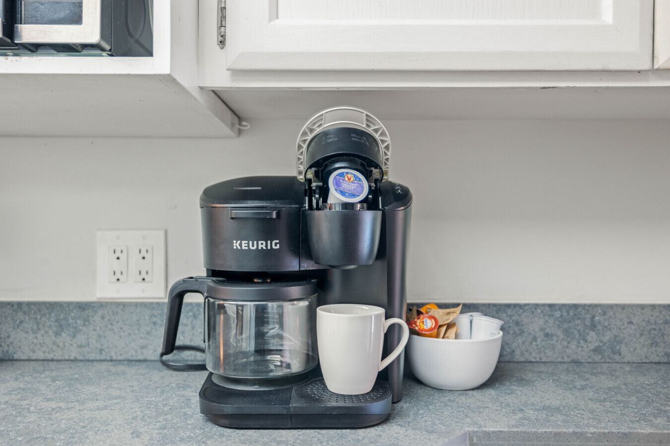 A Keurig coffee maker with a glass carafe, a white mug, and a bowl of coffee pods on a kitchen counter.