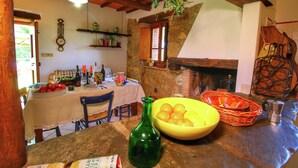 A rustic dining area with a stone wall, wooden beams, a table set with a bowl of fruit, a green bottle, and a basket. - Rustic Holiday Home, Ortignano (Ortignano Raggiolo)