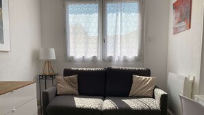 A small, well-lit room with a dark-colored sofa, a small table, and a window with patterned curtains. - Charming T2 Apartment with Private Garage and Balcony in Central Village (Balaruc-les-Bains)